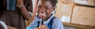 Smiling child sitting at a school desk eating Mary's Meals from a cup