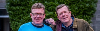 Charlie Reid stands on the left in a Mary's Meals t-shirt holding a mug and Craig Reid rests his arm on Charlie's shoulder and also holds a Mary's Meals mug.