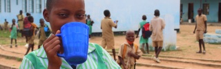 A girl in a green shirt with a blue cup covering her mouth, she is standing outside a school with children behind her