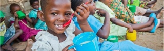 Children in Malawi eating from mugs while sitting on the ground together. The child closest to the camera is looking at it and smiling