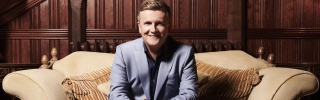 Aled Jones sitting on a sofa with his hands clasped and smiling