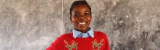 Anna wearing a red Christmas jumper with reindeer on, standing in front of a blackboard smiling