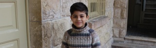 A young boy, Wissam, stands in a doorway smiling. He has dark hair and is wearing a striped top.