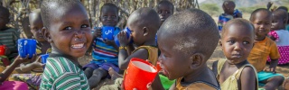 Two children, under 6, from Kenya sitting on the ground eating porridge from mugs