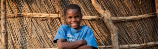 Boy stands in front of home with arms folded and smiling in Zambia