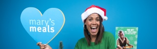 Jean Johansson sitting in a Santa hat, next to a Christmas table, holding up a Mary's Meals sign in one hand and a Christmas gift card in the other