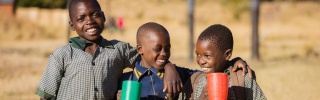 Three children with mugs of Mary's Meals porridge standing outside, smiling, in Zambia