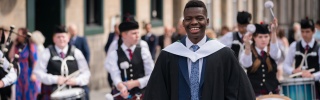 A smiling man in graduation gown stands in front of a Scottish pipe band
