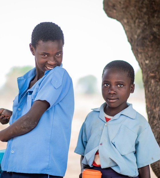Fridah and Annette standing together next to a tree. Fridah is smiling and Annette is holding a mug