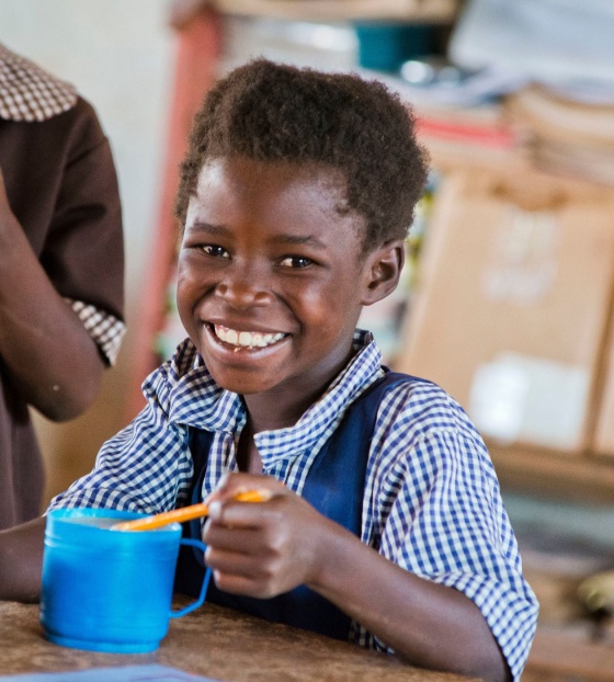 Smiling child sitting at a school desk eating Mary's Meals from a cup