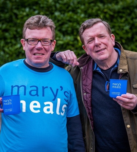 Charlie Reid stands on the left in a Mary's Meals t-shirt holding a mug and Craig Reid rests his arm on Charlie's shoulder and also holds a Mary's Meals mug.