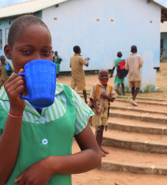 A girl in a green shirt with a blue cup covering her mouth, she is standing outside a school with children behind her