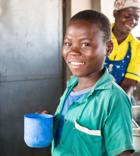 child in kitchen holding a mug, they are smiling. A woman cooks in a large stove behind the child.