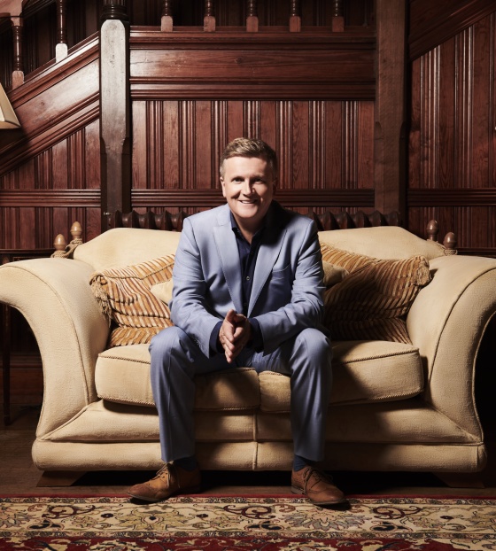 Aled Jones sitting on a sofa with his hands clasped and smiling