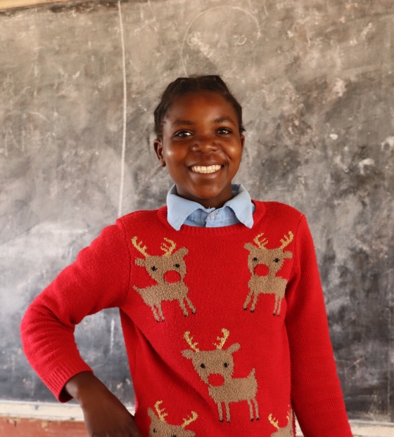 Anna wearing a red Christmas jumper with reindeer on, standing in front of a blackboard smiling