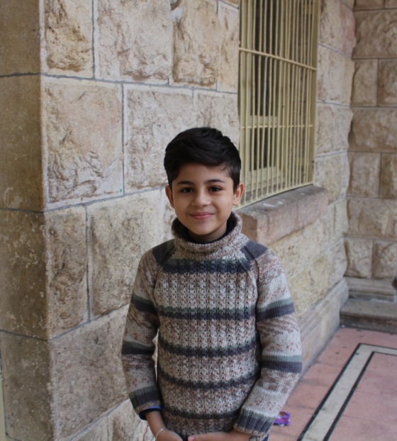 A young boy, Wissam, stands in a doorway smiling. He has dark hair and is wearing a striped top.