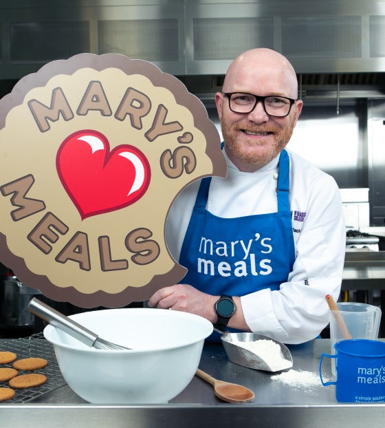 Chef Gary Maclean with Mary's Meals blue apron on and biscuit prop