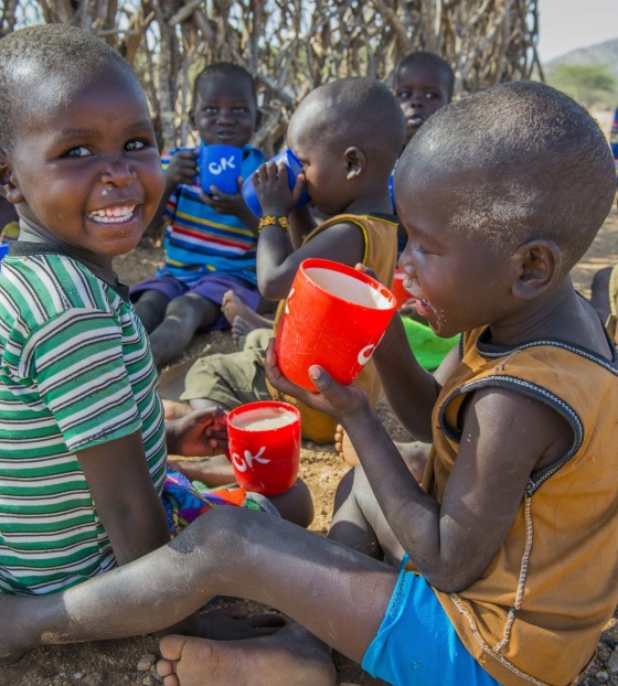 Two children, under 6, from Kenya sitting on the ground eating porridge from mugs