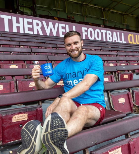 Football Stephen O'Donnell sitting in stand of football stadium in blue Mary's Meals t-shirt with blue Mary's Meals mug