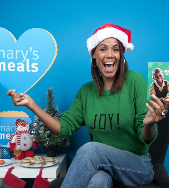 Jean Johansson sitting in a Santa hat, next to a Christmas table, holding up a Mary's Meals sign in one hand and a Christmas gift card in the other