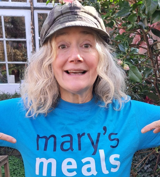 Actor Sophie Thompson stands outside wearing a Mary's Meals t-shirt, she is smiling and pointing at the Mary's Meals logo on the t-shirt
