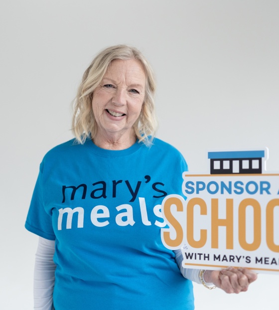 Deborah Meaden wearing a Mary's Meals t-shirt holding a sign which reads 'Sponsor A School with Mary's Meals'
