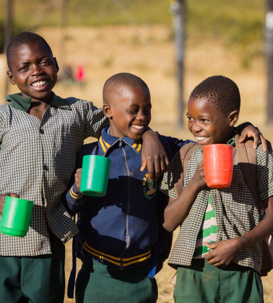 Three children with mugs of Mary's Meals porridge standing outside, smiling, in Zambia