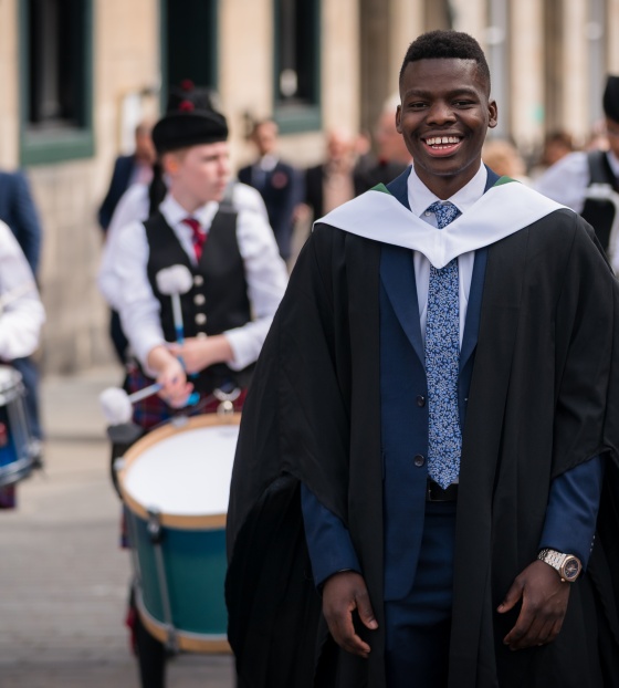A smiling man in graduation gown stands in front of a Scottish pipe band