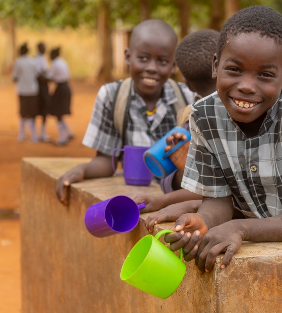 Children smiling while leaning on wall with mugs of Mary's Meals porridge in Zambia