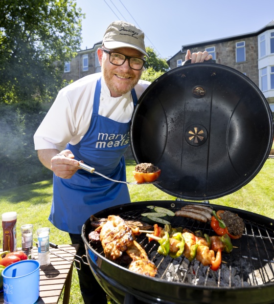 Gary Maclean holds a piece of meat over a BBQ with food cooking