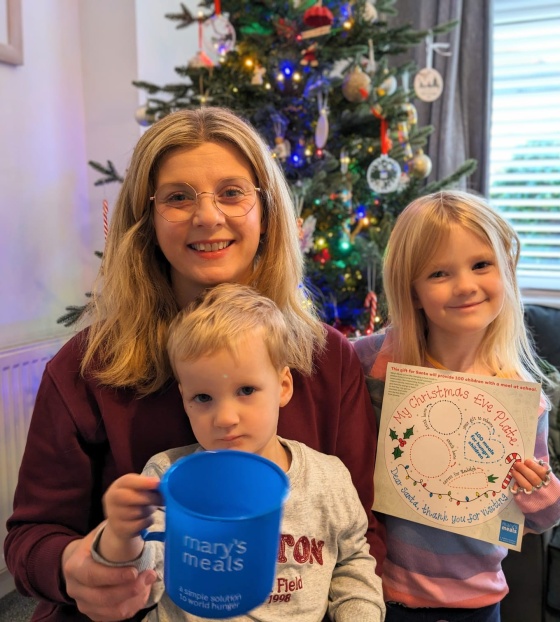 Victoria and her children in a living room, a boy holds a Mary's Meals mug and a girl holds a Santa plate
