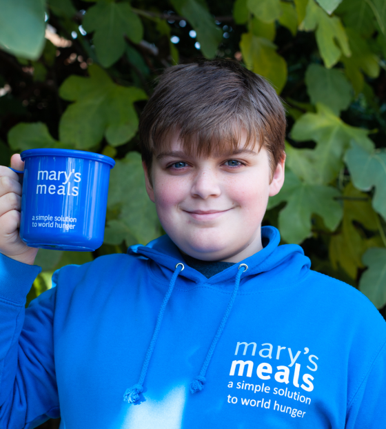 A young boy stands in a Mary's Meals hoodie holding a Mary's Meals mug