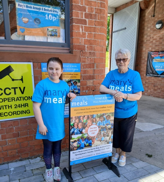 A woman and a girl stand in Mary's Meals t-shirts next to a Mary's Meals sign