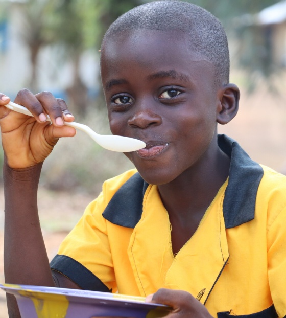 A boy in yellow sits outside eating a bowl of food with a spoon