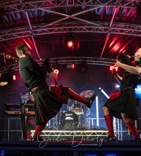 A group of men in kilts play bagpipes and guitars on a stage
