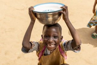 A young child from Benin carrying a silver bowl on his head and smiling at the camera