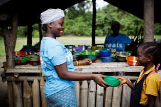 A volunteer cook is handing over a bowl of porridge to a child