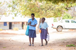 Fridah and Annette walking together outside. They are carrying bags.