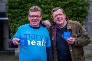 Charlie Reid stands on the left in a Mary's Meals t-shirt holding a mug and Craig Reid rests his arm on Charlie's shoulder and also holds a Mary's Meals mug.