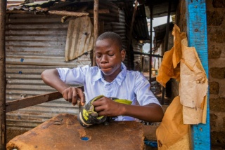 A teenager sits at a small desk repairing a pair of old shoes