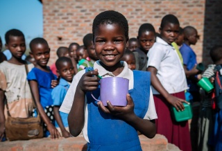 Smiling girl with mug and queue of children waiting to be served Mary's Meals behind her