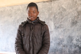 Schoolboy pictured wearing jacket in classroom in front of blackboard in Malawi