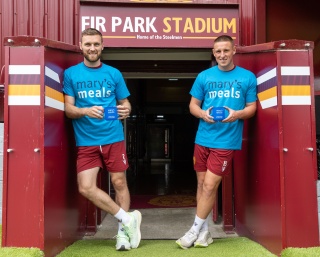 Motherwell footballers Stephen O'Donnell and Dan Casey stand beside each other at Fir Park Stadium tunnel in blue Mary's Meals t-shirts holding blue Mary's Meals mugs