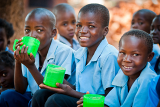 Boys eating Mary's Meals from green cups, in blue shirts, looking to camera