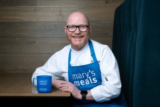 Gary Maclean sits at a table which holds a Mary's Meals mug. He's smiling and wearing a Mary's Meals apron.