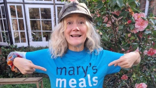 Actor Sophie Thompson stands outside wearing a Mary's Meals t-shirt, she is smiling and pointing at the Mary's Meals logo on the t-shirt
