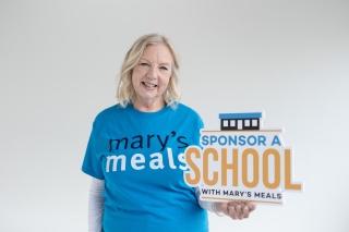 Deborah Meaden wearing a Mary's Meals t-shirt holding a sign which reads 'Sponsor A School with Mary's Meals'