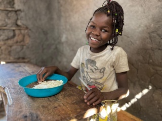 A girl in Mozambique smiles while she sits at a table with a bowl of food