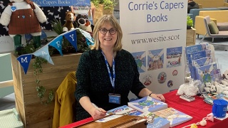 A lady, Alison Page, sits at a stall selling her children's books