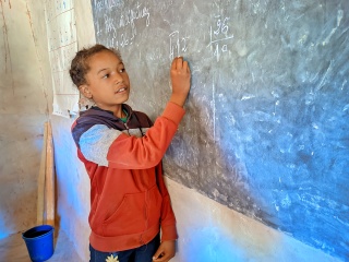 A young girl, Melanie, writes on a blackboard in a classroom